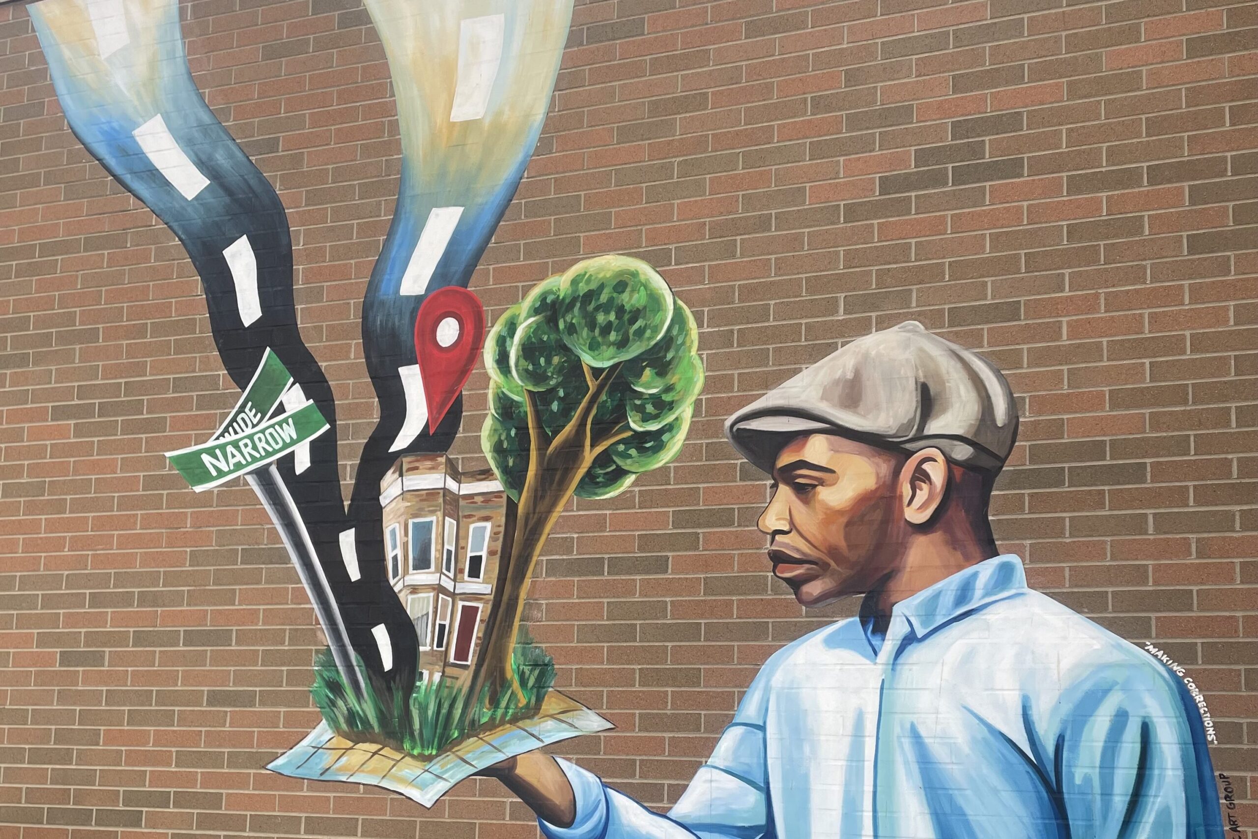 Art painted on a brick wall. On the right stands a Black man in a cap and a blue shirt. He is facing left with a solemn expression and extending his hand. In his hand is a map of a city block. Emerging from the map is a house, a tree, two roads, and a street sign that says WIDE and NARROW.